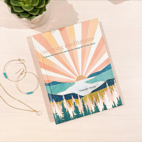 A small hard back book called 'Morning Meditations', with a peach, pink and white sunburst design over turquoise and teal hills with green, pink and white trees in the foreground.