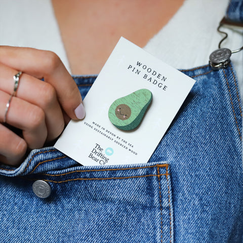 Wooded pin badge in the shape of an avocado with cute smiley face. Printed on walnut veneer, pinned onto a recycled backing card which reads 'Wooden Pin Badge' in large text and 'Made in Devon by the sea using sustainably sourced wood' in small text.