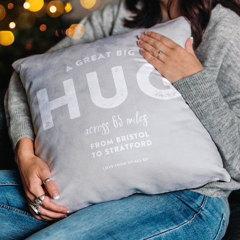 A woman sitting in front of a Christmas tree, holds a luxurious, plump cushion. The cushion has white text on a light grey background that reads 'A great big hug across 65 miles from Bristol to Stratford, love from us all xx. 