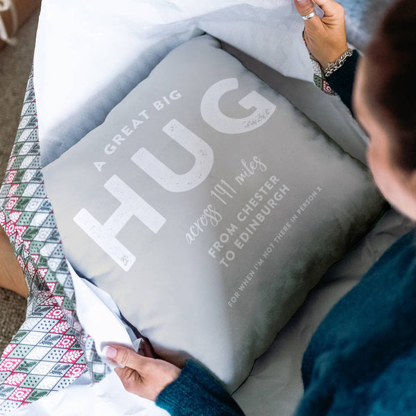 The image shows a woman unwrapping a Christmas present, inside is a personalised cushion stating the words ' a great big hug across 191 miles from Chester to Edinburgh, for when I'm not there in person x'.